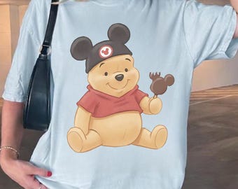 May include: Light blue t-shirt featuring a cartoon illustration of Winnie the Pooh wearing a Mickey Mouse hat and holding a Mickey Mouse-shaped ice cream bar. The bear is wearing a red shirt. The shirt is a light blue color.