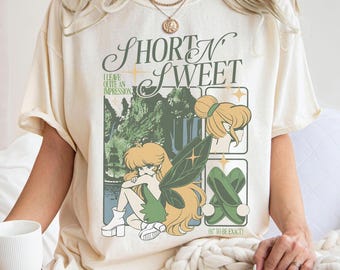 May include: Cream-colored t-shirt featuring a graphic with the text "Short N Sweet" and an illustration of a fairy. The fairy has blonde hair, green wings, and is wearing green clothing. The shirt also has the text "I leave quite an impression."