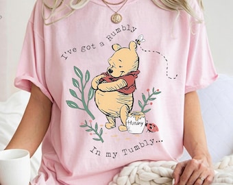May include: Pink t-shirt featuring a graphic of Winnie the Pooh with the text "I've got a Rumbly in my Tumbly...". The design includes a honey pot, a bee, and foliage. The shirt has short sleeves.