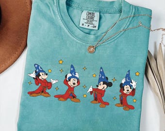 May include: Teal Comfort Colors t-shirt featuring a graphic of Mickey Mouse in a sorcerer's outfit. The design includes Mickey in a red robe and blue wizard hat, with yellow stars. A gold necklace is draped across the shirt.
