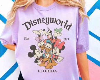 May include: A light purple t-shirt featuring a graphic of Disney characters including Mickey Mouse, Minnie Mouse, Donald Duck, Daisy Duck, Goofy, and Pluto. The text "Disneyworld" is arched above the characters, with "Est 1971" and "Florida" below.