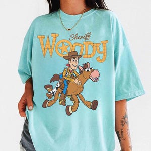 May include: A light blue t-shirt featuring a graphic of Sheriff Woody riding a horse. The text "Sheriff Woody" is printed above the graphic in orange and yellow. The shirt has short sleeves and a crew neck.