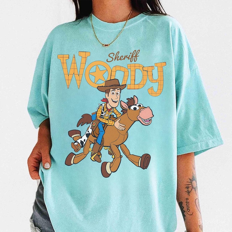 May include: A light blue t-shirt featuring a graphic of Sheriff Woody riding a horse. The text "Sheriff Woody" is printed above the graphic in orange and yellow. The shirt has short sleeves and a crew neck.