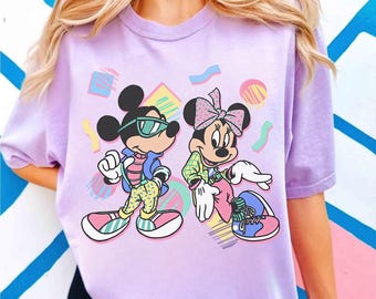 May include: A light purple t-shirt featuring a cartoon illustration of Mickey and Minnie Mouse in 80s-inspired outfits. Mickey wears sunglasses, a jacket, and patterned pants. Minnie has a bow and a plaid shirt. Geometric shapes surround them.