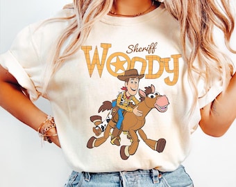 May include: Cream-colored t-shirt featuring a cartoon illustration of Sheriff Woody riding a horse. The text "Sheriff Woody" is printed above the image in a western-style font. The shirt is made of a soft material.