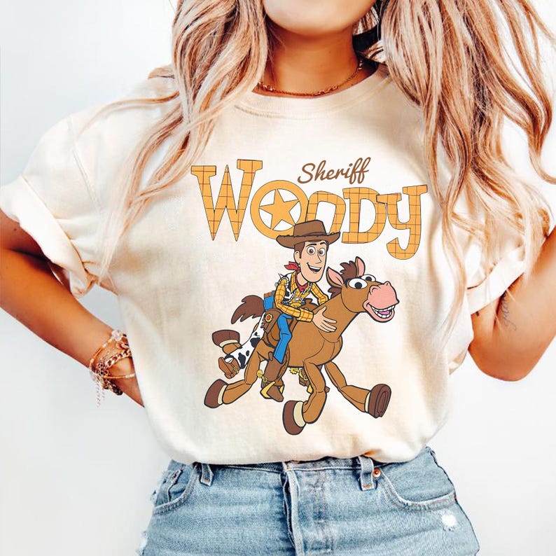 May include: Cream-colored t-shirt featuring a cartoon illustration of Sheriff Woody riding a horse. The text "Sheriff Woody" is printed above the image in a western-style font. The shirt is made of a soft material.