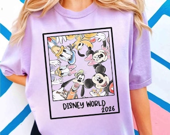 May include: A light purple t-shirt featuring a framed graphic of Disney characters, including Mickey Mouse, Minnie Mouse, Donald Duck, and Goofy. The graphic is labeled "DISNEY WORLD 2026".