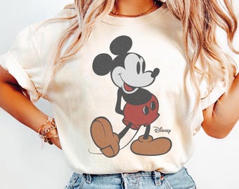 May include: Cream-colored t-shirt featuring a classic Mickey Mouse graphic. The cartoon character is depicted in black, red, and brown, with the Disney logo printed below. The shirt is paired with blue denim jeans.