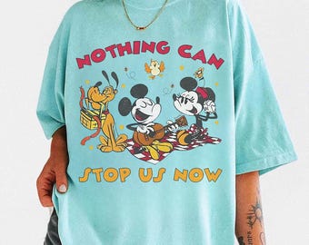 May include: A light blue t-shirt featuring a graphic of Mickey and Minnie Mouse playing guitar on a picnic blanket, with Pluto. The text "NOTHING CAN STOP US NOW" is printed above and below the characters.