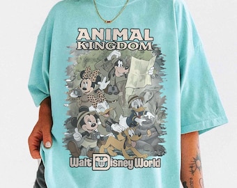 May include: A light blue t-shirt featuring a graphic of Disney characters, including Mickey Mouse, Minnie Mouse, Donald Duck, and Goofy, in safari attire. The shirt has the words "ANIMAL KINGDOM" and "Walt Disney World" printed on it.
