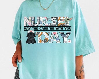 May include: A light blue t-shirt with the words "NURSE DAY" in a block letter design. The text "MAY THE CARE BE WITH YOU" is printed between the words. The letters are filled with medical and Star Wars themed graphics.