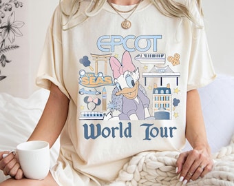 May include: Cream-colored t-shirt featuring Daisy Duck in a purple outfit with a pink bow. The shirt has the text "EPCOT World Tour" in blue, along with various travel-themed illustrations. The shirt is a comfortable, casual style.