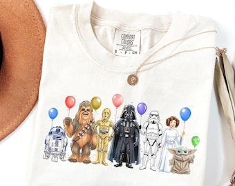 May include: Cream-colored t-shirt featuring a graphic of Star Wars characters holding balloons. Characters include R2-D2, Chewbacca, C-3PO, Darth Vader, a Stormtrooper, Princess Leia, and Grogu. The shirt has a Comfort Colors tag.