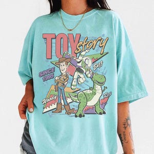 May include: A light blue t-shirt featuring the words "TOY Story" in pink and a graphic of Woody, Buzz Lightyear, and Rex. The shirt also includes the text "SINCE 1995".