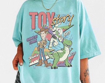 May include: A light blue t-shirt featuring the words "TOY Story" in pink and a graphic of Woody, Buzz Lightyear, and Rex. The shirt also includes the text "SINCE 1995".