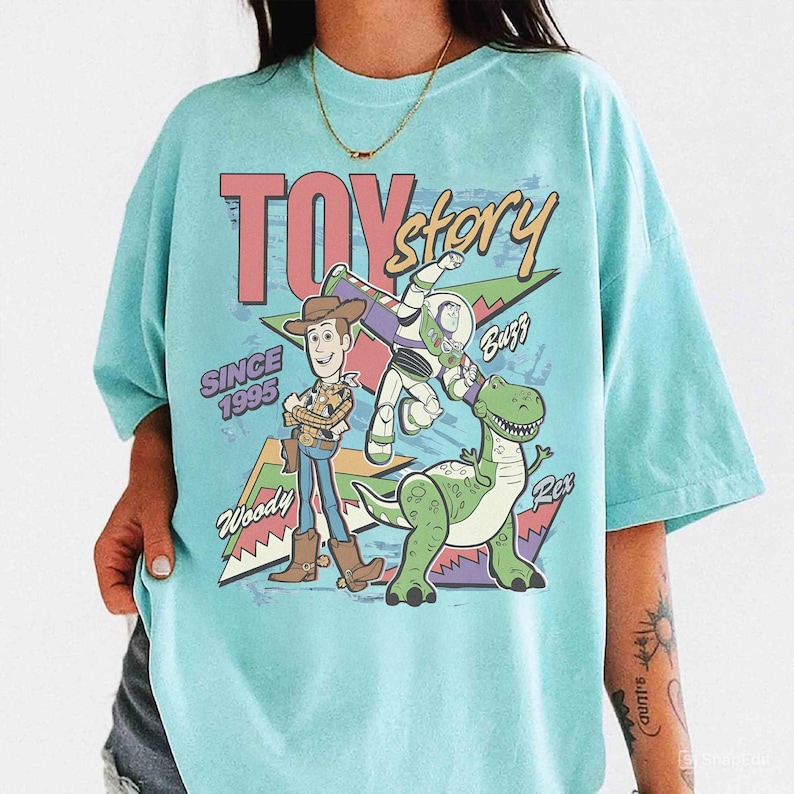 May include: A light blue t-shirt featuring the words "TOY Story" in pink and a graphic of Woody, Buzz Lightyear, and Rex. The shirt also includes the text "SINCE 1995".