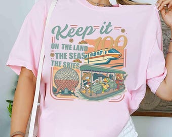 May include: A light pink t-shirt featuring the text "Keep it 100" with illustrations of a train, a boat with cartoon characters, and a geodesic sphere. The shirt has short sleeves and a crew neck.