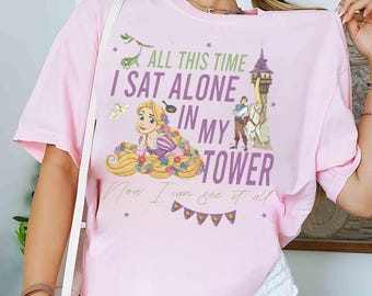 May include: A light pink t-shirt featuring a graphic of a princess with long blonde hair, flowers, and a tower. The text on the shirt reads "ALL THIS TIME I SAT ALONE IN MY TOWER Now I can see it all."
