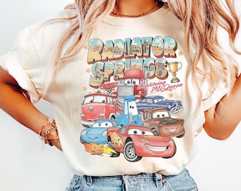May include: Cream-colored t-shirt featuring a colorful graphic of characters from the movie Cars, including Lightning McQueen and the text "Radiator Springs". The design includes a trophy and stars.