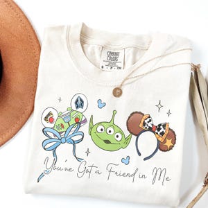 May include: Cream-colored t-shirt featuring cartoon characters and the text "You've Got a Friend in Me." The design includes Buzz Lightyear, a green alien, and mouse ears. A gold necklace and a brown hat are also visible.