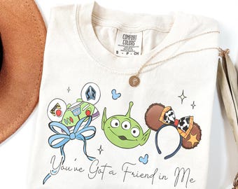 May include: Cream-colored t-shirt featuring cartoon characters and the text "You've Got a Friend in Me." The design includes Buzz Lightyear, a green alien, and mouse ears. A gold necklace and a brown hat are also visible.
