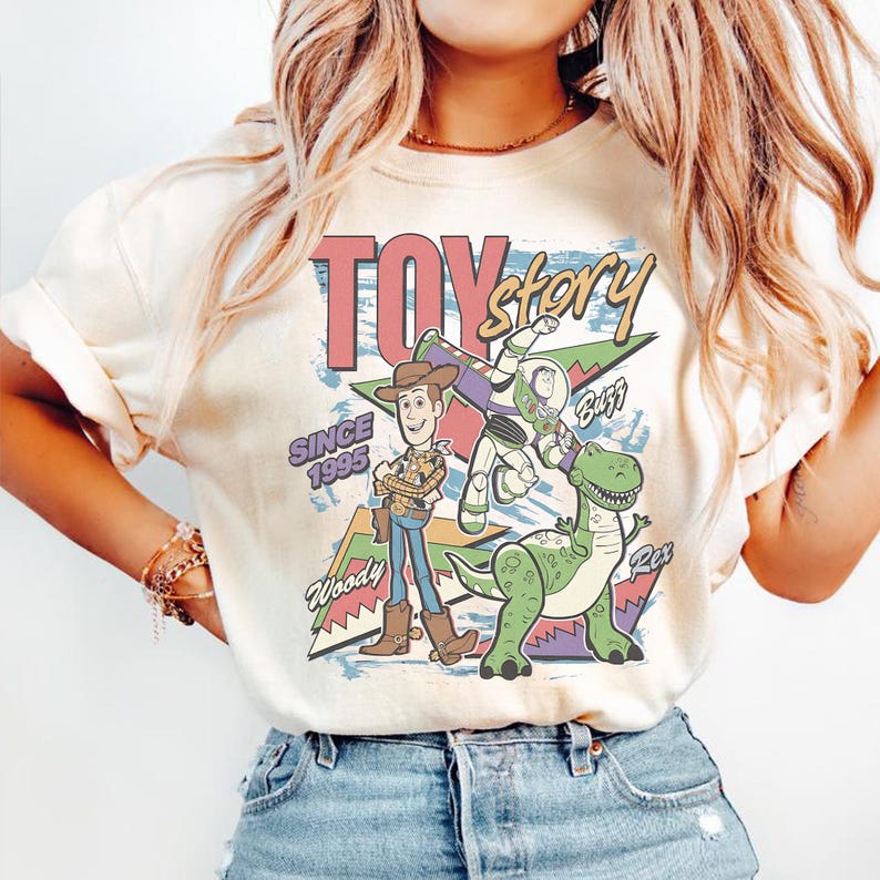 May include: Cream-colored t-shirt featuring a graphic of Toy Story characters Woody, Buzz Lightyear, and Rex, with the text "TOY STORY" and "SINCE 1995". The design includes a retro, cartoon-style aesthetic.