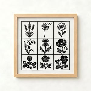 May include: A square framed cross-stitch embroidery featuring a grid of nine distinct black botanical illustrations on a white fabric background. The designs include various wildflowers and plants such as lavender, a dandelion, and a poppy.