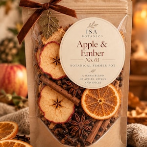 Op de afbeelding: Een kraftpapieren zakje met een botanische sudderpotmix, gelabeld ISA Botanics Apple & Ember No. 01. Het doorzichtige venster toont gedroogde appelschijfjes, sinaasappelschijfjes, kaneelstokjes, steranijs en kruidnagels. Een bruin fluwelen lint en een metalen bladbedel zijn bevestigd.