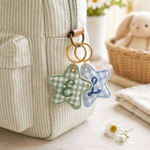 May include: Two star-shaped keyrings with embroidered letters, "E" and "L", hanging from a bag. One keyring is green gingham, the other is blue gingham. The bag is striped in white and light green.