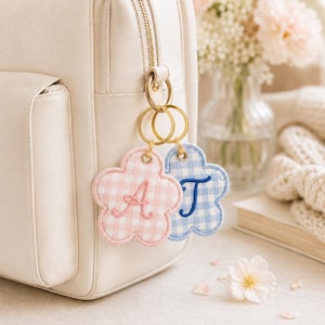 Puede incluir: Dos llaveros de tela con forma de flor sujetos a la cremallera de una mochila color crema. Uno tiene un patrón de cuadros vichy rosa con la letra A bordada, y el otro un patrón de cuadros vichy azul con la letra T bordada.