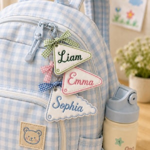 May include: A light blue and white gingham backpack featuring a bear patch and three triangular personalized keychains with the names Liam, Emma, and Sophia, each accented with a small fabric bow attached to the zipper pull of the bag.