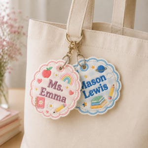 Puede incluir: Dos etiquetas bordadas de borde festoneado para bolso, sujetas a un bolso de lona color crema. Una etiqueta es rosa claro con el nombre Ms. Emma, una manzana, un arcoíris y un lápiz. La otra es azul claro con el nombre Mason Lewis, un planeta y libros.