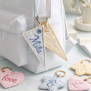 May include: A light blue and white striped backpack with gold-colored hardware. Attached are two pennant-shaped keychains, one blue with "Mila" embroidered, and one yellow with "Clara". Other keychains include heart and star shapes.