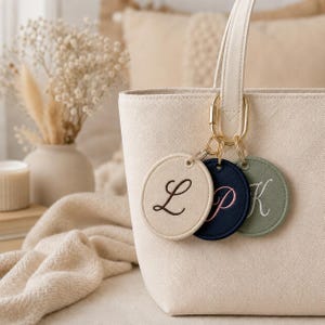 May include: A cream-coloured canvas tote bag featuring three circular fabric keychains attached to the handle. The keychains are beige with a dark brown letter L, navy blue with a pink letter P, and sage green with a white letter K embroidered.