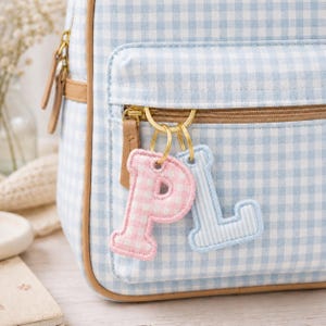 May include: A light blue gingham backpack with a gold zipper and leather accents. Two letter keychains, a pink "P" and a blue "L", hang from the zipper. The keychains have a stitched border.