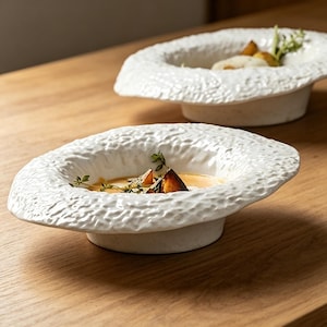 May include: Two white ceramic soup bowls with a textured rim and a shallow base. Each bowl contains soup and garnishes. The bowls are on a wooden surface, creating a warm, inviting aesthetic.