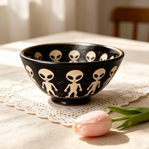 Peut inclure: Un bol en céramique noire orné d'un motif répétitif de personnages extraterrestres couleur crème, dotés de grands yeux ovales et de corps fins. Le motif est imprimé sur l'extérieur et le bord intérieur du bol, qui présente une finition mate.