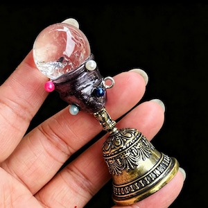 May include: A decorative object featuring a clear spherical crystal orb set into a dark, textured, cone-shaped base adorned with small white, pink, and blue beads. The base is attached to a metallic gold-toned bell with intricate embossed patterns.