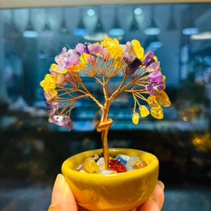 Puede incluir: Una escultura decorativa de árbol con tronco y ramas de alambre de cobre, adornada con hojas de piedras preciosas de amatista y amarillas. El árbol está en una maceta de cerámica amarilla llena de piedras de colores. La escultura se sostiene en una mano.