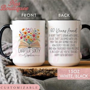 May include: A 15oz white ceramic mug with a black handle and rim. The front features an open book with colorful wildflowers and the text Chapter Sixty Sophia. The back displays the text 60 Years Loved with a heartfelt birthday message.