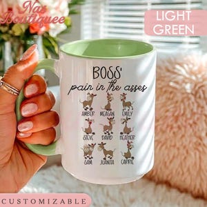 May include: A white ceramic mug with a light green interior and handle. The mug features the black text 'BOSS' pain in the asses' above nine illustrations of donkeys, each labeled with a name: Amber, Meagan, Emily, Steve, David, Heather, Sam, Juanita, and Carrie.