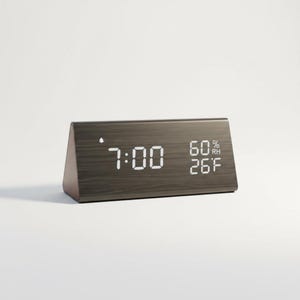 May include: A digital alarm clock with a dark grey, wood-grain finish. The display shows the time as 7:00, humidity at 60%, and the temperature at 26 degrees Fahrenheit. The clock has a triangular shape.