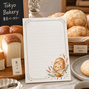 May include: A rectangular notepad with horizontal lines and a decorative border. The bottom right corner features a cartoon illustration of a smiling bread loaf wearing a checkered scarf, surrounded by wheat stalks and small star accents.