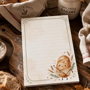 May include: A rectangular notepad with horizontal lines and a rounded border in muted green. The bottom right corner features a cartoon illustration of a smiling loaf of bread wearing a checkered scarf, surrounded by wheat stalks and small stars.