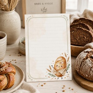 May include: A rectangular stationery sheet with horizontal lines, featuring a rounded green border and a watercolor illustration of a smiling anthropomorphic loaf of bread wearing a checkered scarf, surrounded by wheat stalks and small green leaves.