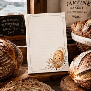 May include: A rectangular notepad with horizontal lines and a decorative border. The bottom right corner features a cartoon illustration of a smiling bread loaf wearing a checkered apron, surrounded by small wheat stalks and star accents.
