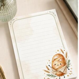 May include: A rectangular notepad featuring horizontal lines and a decorative border in muted green tones. The bottom right corner displays a whimsical illustration of a smiling loaf of bread wrapped in a checkered cloth, surrounded by wheat stalks.