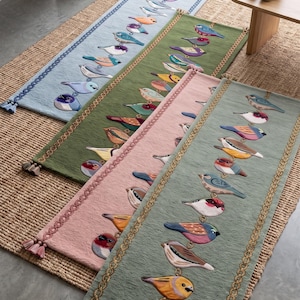 May include: Four rectangular table runners in light blue, olive green, dusty pink, and sage green. Each runner features a vertical row of embroidered colorful birds, decorative braided side borders, and matching tassels at the bottom corners.