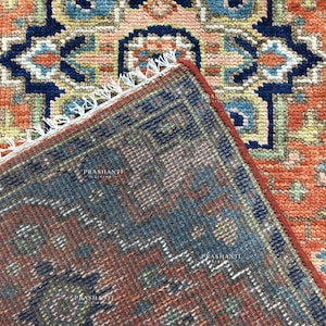 K&ouml;nnte beinhalten: Nahaufnahme eines Teppichabschnitts mit detailliertem, geometrischem Muster. Der Teppich weist eine Farbkombination aus Orange, Marineblau und Akzenten in Hellblau und Beige auf. Der Rand des Teppichs ist sichtbar und zeigt einen Fransenbesatz.