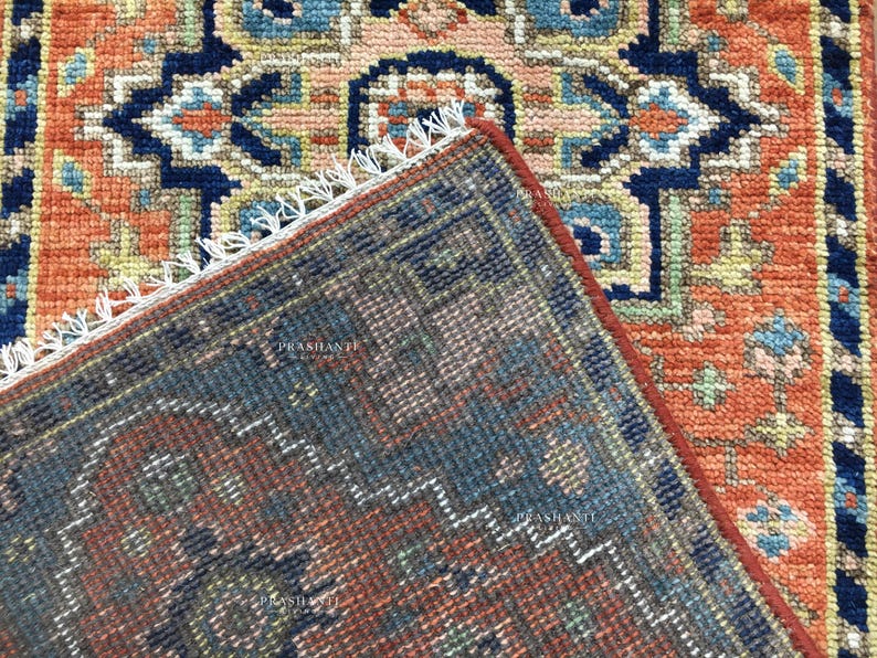 K&ouml;nnte beinhalten: Nahaufnahme eines Teppichabschnitts mit detailliertem, geometrischem Muster. Der Teppich weist eine Farbkombination aus Orange, Marineblau und Akzenten in Hellblau und Beige auf. Der Rand des Teppichs ist sichtbar und zeigt einen Fransenbesatz.
